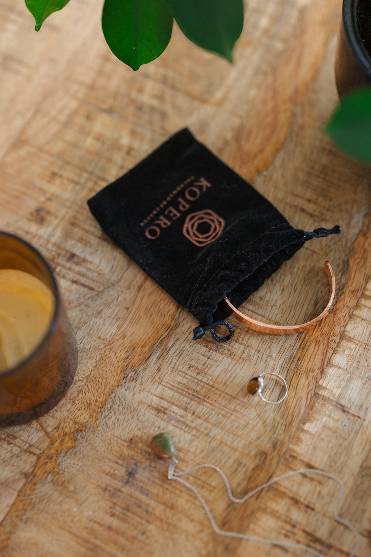 Kopero Kupferarmband Glatt, auf einem Holztisch neben dem Samtbeutel, einer Kerze und Schmuck