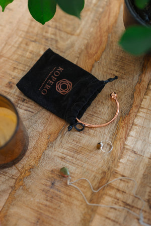 Kopero Kupferarmband Rund, auf einem Holztisch neben dem Samtbeutel, einer Kerze und Schmuck