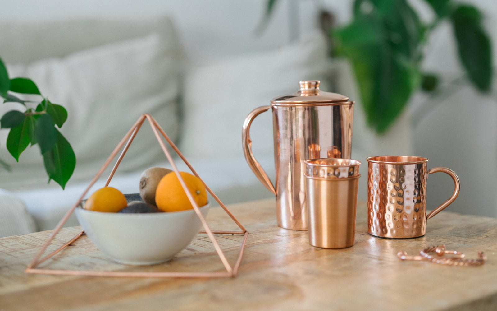 Kupferpyramide über einer Obstschale, daneben eine Kanne, ein Becher, eine Tasse und ein Armband auf einem Holztisch