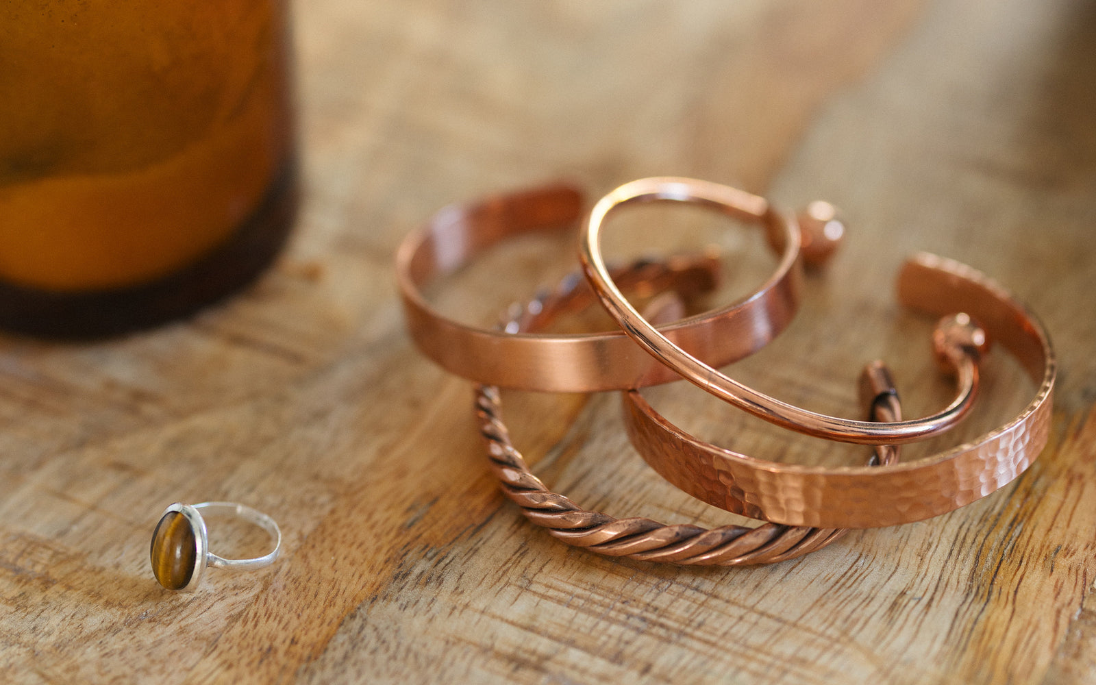 Vier Kupferarmbänder, gedreht, glatt, gehämmert und rund, auf einem Holztisch neben einer Kerze und einem Ring