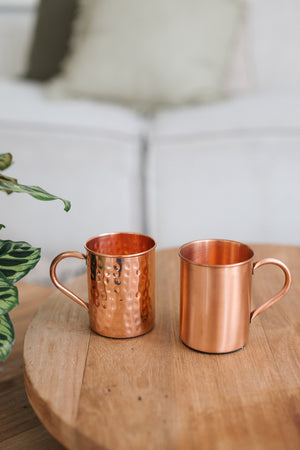 Kopero Kupfertasse, eine glatte und eine gehämmerte Tasse nebeneinander auf einem runden Holztisch