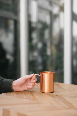 Kopero Kupfertasse Glatt, auf einem Holztisch, mit einer Hand am Henkel