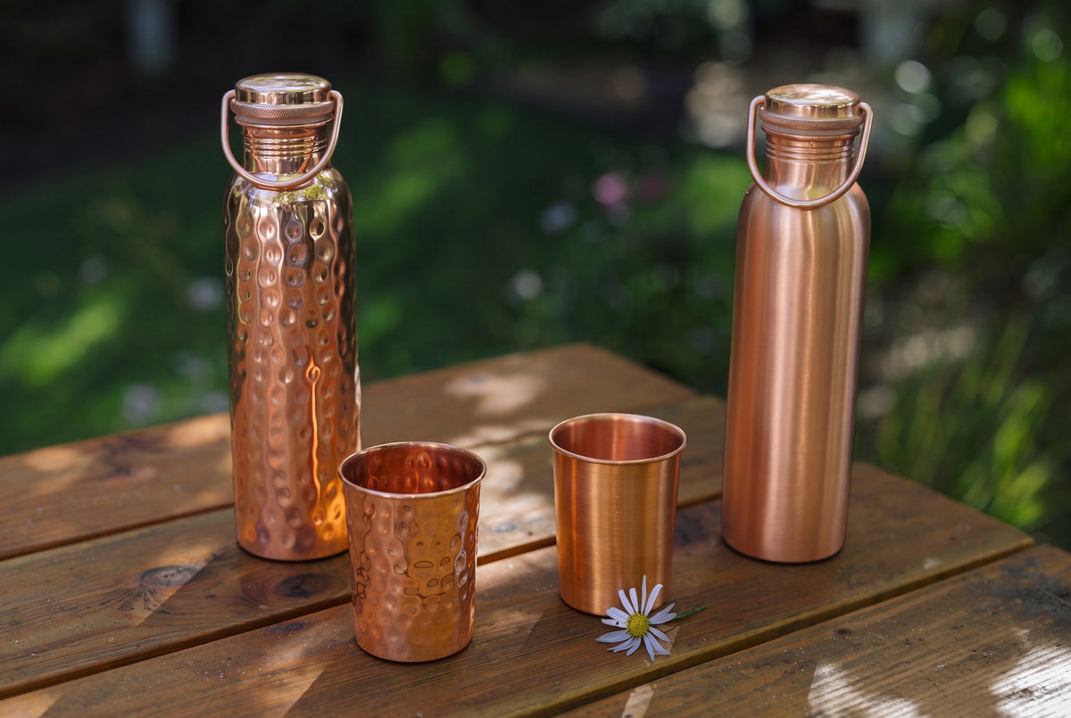 Kupferflaschen, glatte und gehämmerte Becher und ein Gänseblümchen auf einem Holztisch im Garten